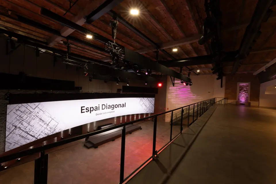 Large curved screen showing the Espai Diagonal sign, seen from the gallery, with a low stage and black ceiling beams