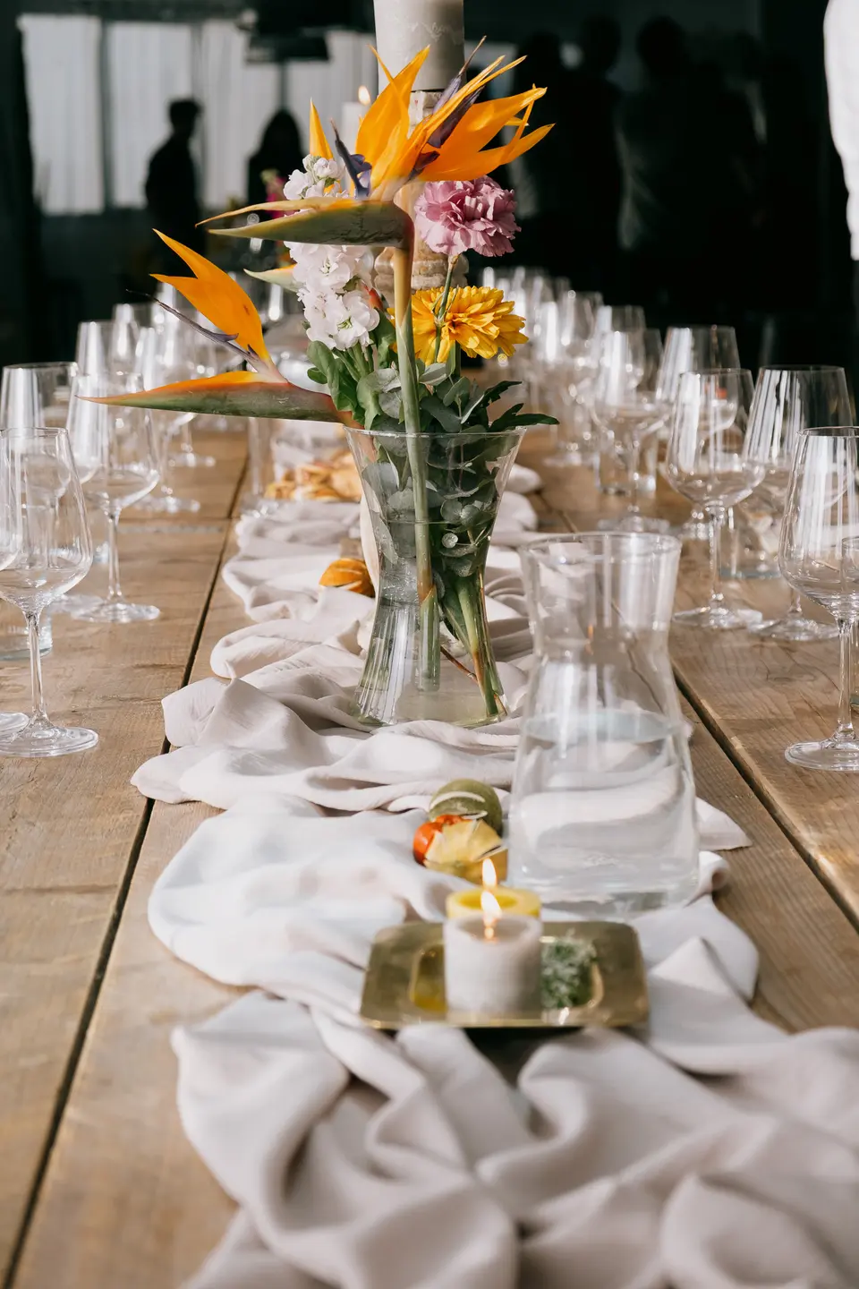 Wooden table with a beige fabric runner, glasses, a vase of orange tropical flowers and a lit candle