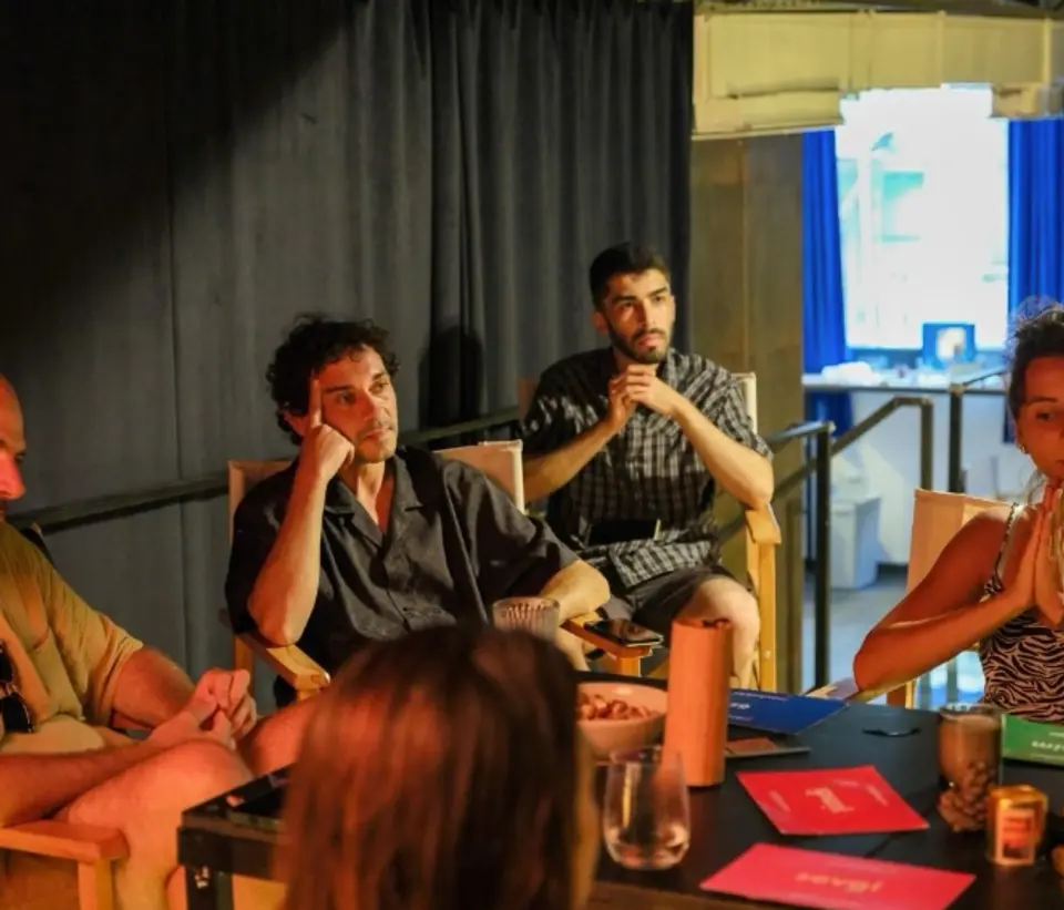 Several people seated in wooden chairs around a table with red cards, listening attentively