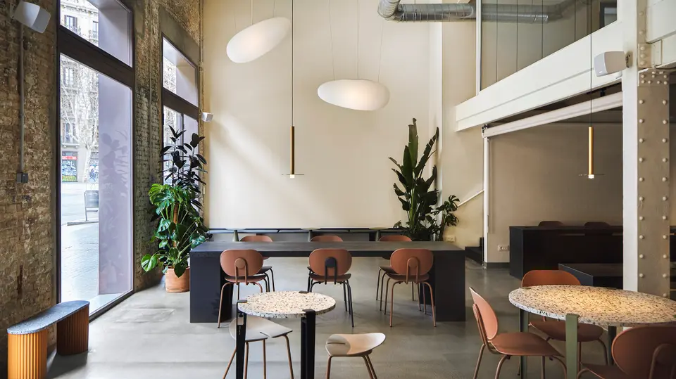 Double-height room with a black table, terracotta chairs, terrazzo tables and large windows facing the street