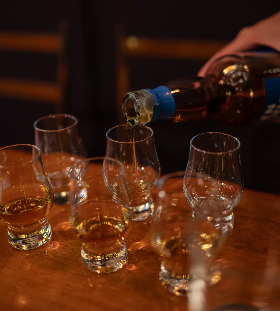 Hand pouring whisky from a bottle into a row of tasting glasses on a wooden table
