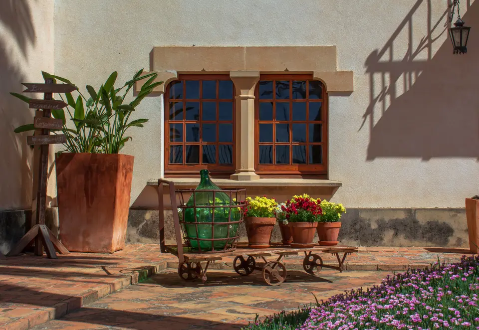 Wooden windows in a pale wall with a tall terracotta pot, colorful flowers and a green demijohn on a cart