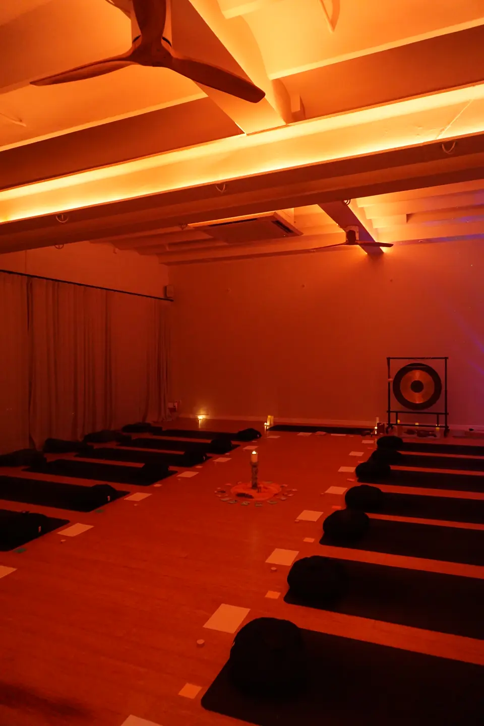 Wide view of the room with rows of mats and cushions leading to a gong, under a warmly lit ceiling