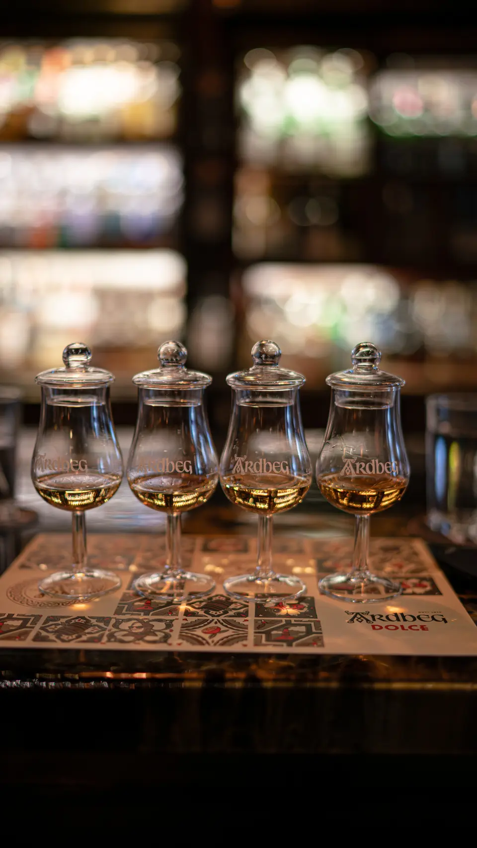 Four lidded tasting glasses lined up on a tiled mat, with shelves of bottles behind them