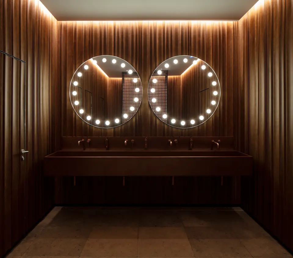 Restroom with dark wood panelling, two round mirrors ringed with bulbs and a long trough sink with five taps