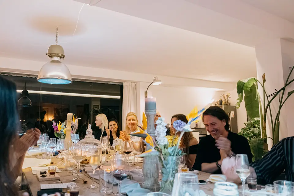 Diners at a long table applauding and smiling, with flowers and candles and a large window behind
