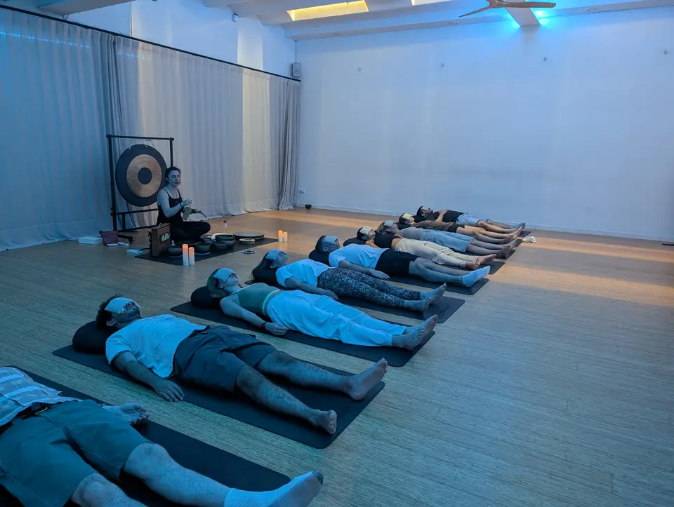 People lying on their backs on mats in a large room, with one person seated beside a gong and candles