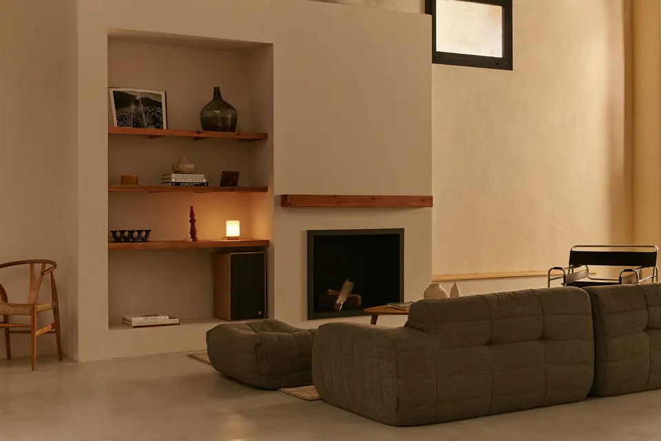 Fireplace corner with wooden shelves and a lit lamp, with a grey sofa seen from behind and a wicker-seat chair