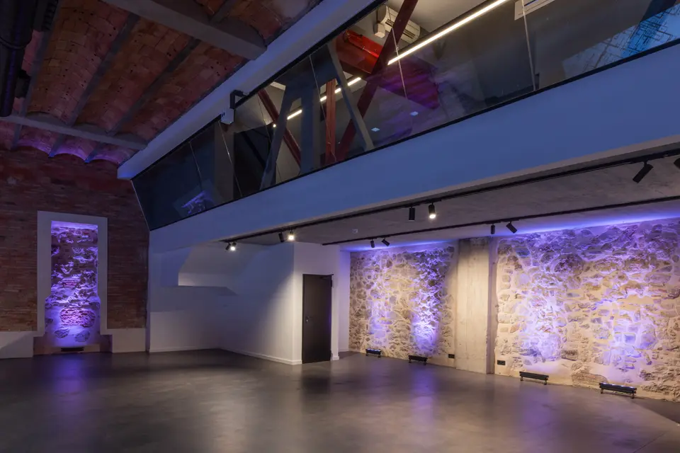 Open hall with a dark floor, stone walls lit in purple and a glass-fronted gallery on the upper level