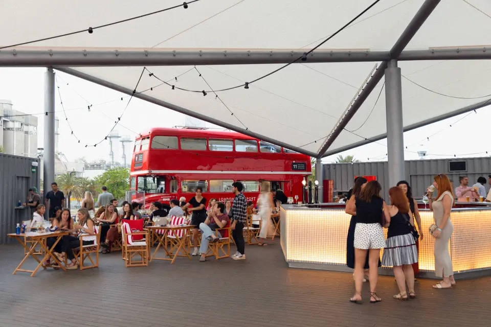 Terrace under a tensioned canopy with a lit bar on the right, wooden tables and a red double-decker bus at the back