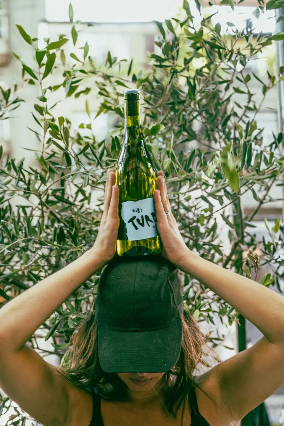 Person in a cap holding a wine bottle above their head in front of olive branches