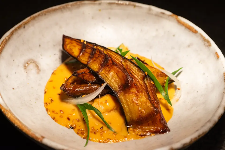 Shallow white ceramic bowl with roasted eggplant over an orange sauce with sesame seeds and spring onion