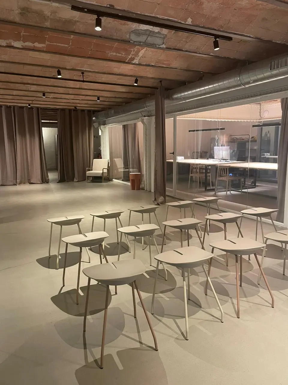 Beige stools set in rows in a theatre-style layout, open room with a polished floor, fabric curtains and a brick ceiling