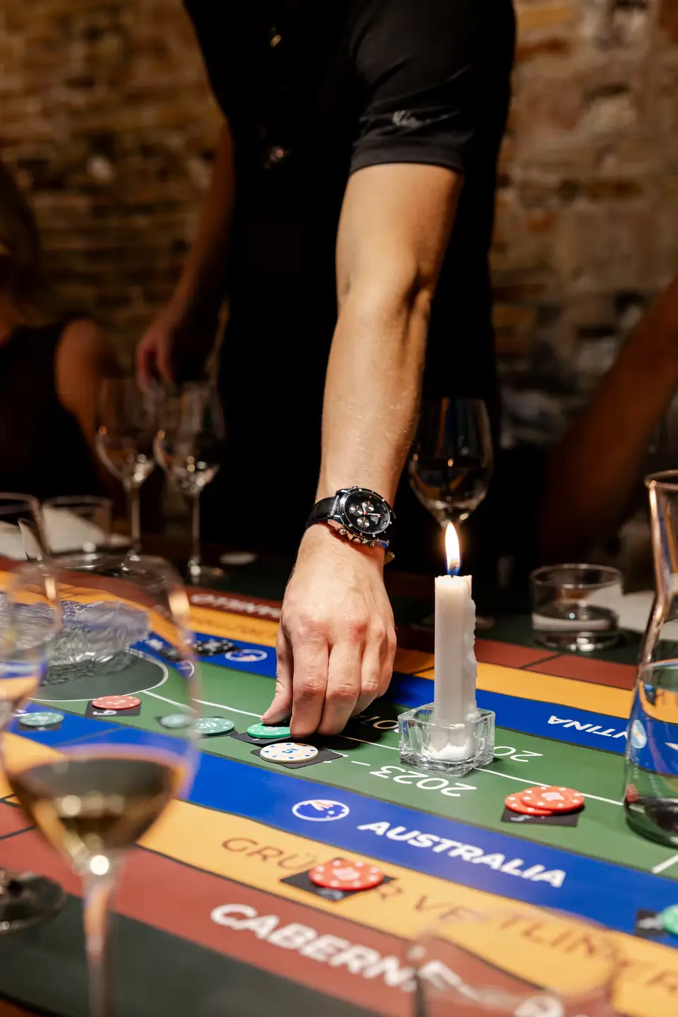 Hand resting on a game board with colored chips, with a lit candle and wine glasses in front
