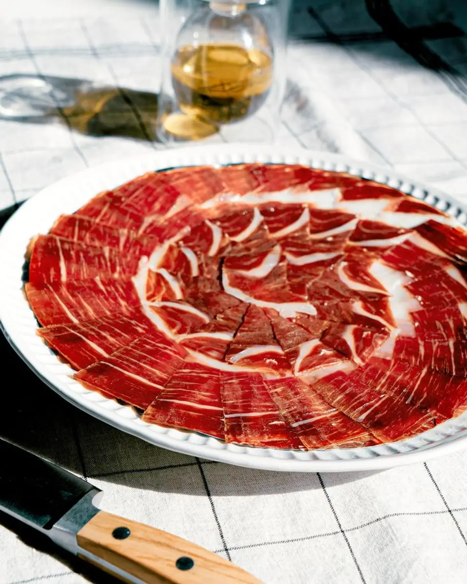 White plate of thinly sliced Iberian ham on a checked tablecloth, with a carving knife beside it