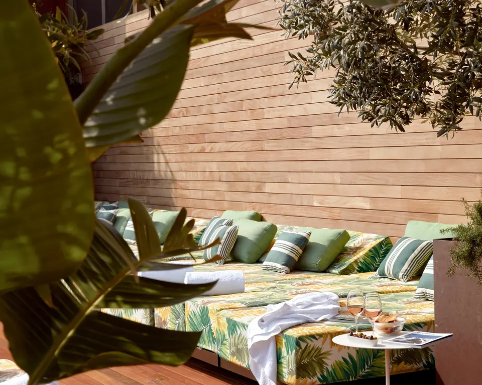 Outdoor daybed with green striped cushions and tropical leaf fabric, next to a wooden wall and an olive tree