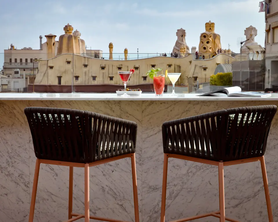 Two woven stools at a marble bar with cocktails and a view of the chimneys of Casa Milà