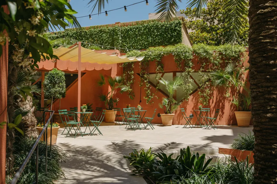 Garden courtyard with green tables and chairs, terracotta walls covered in climbing plants and a yellow awning