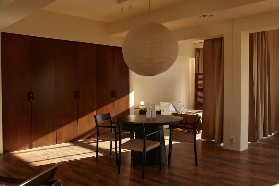 Round black table with three chairs under a paper lamp, beside wooden wardrobes and a bed behind the curtains