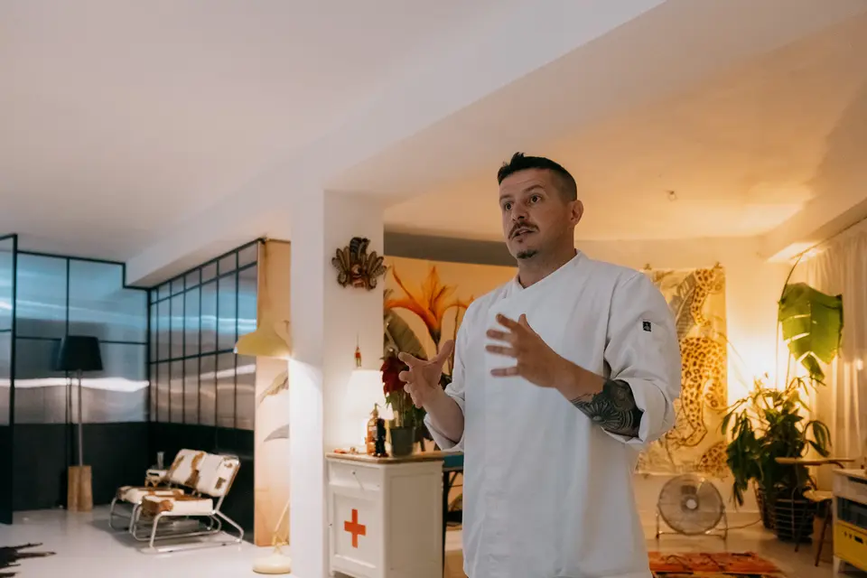 Chef in a white jacket speaking with hand gestures in a white-walled loft, with plants and an armchair behind him