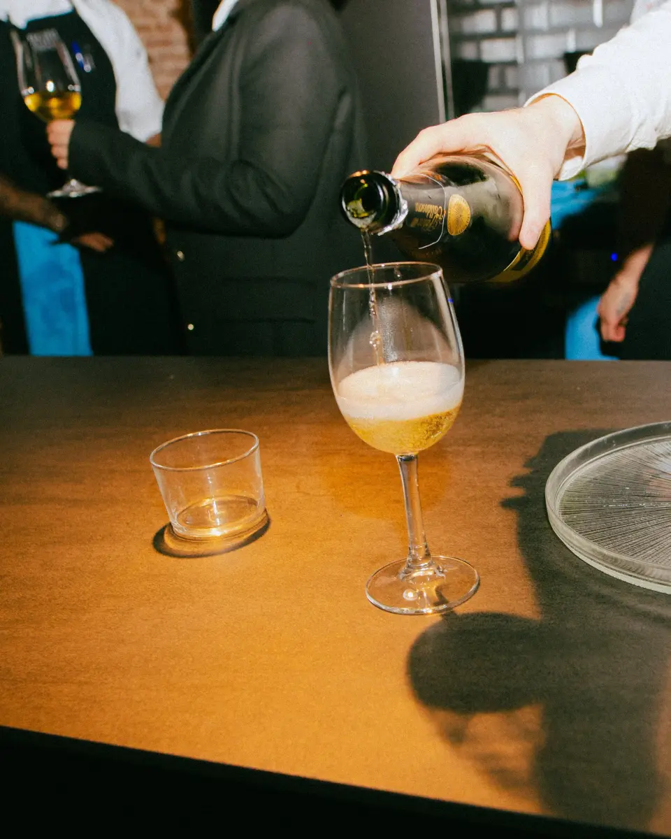 Hand pouring sparkling wine from a bottle into a glass on an amber-colored table