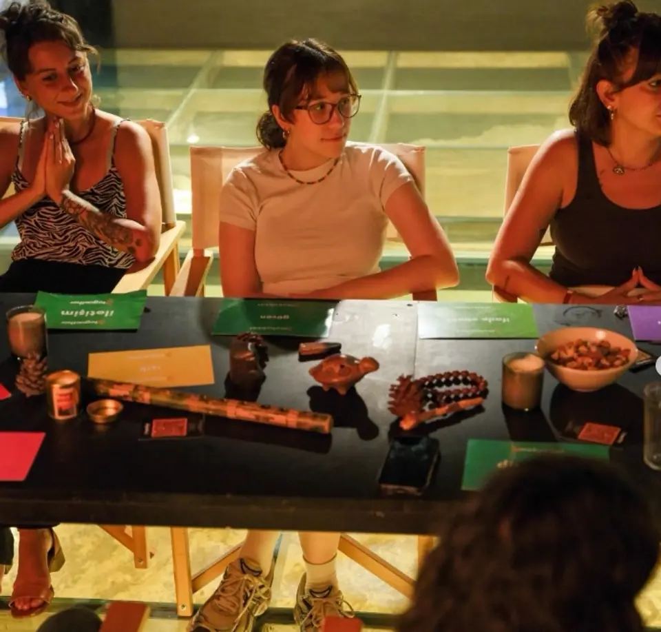 Three people seated at a dark table with green cards, bead necklaces and a bowl of nuts