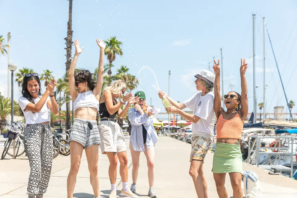 Group with arms raised on a marina quay as foam sprays from a bottle, with palm trees and masts behind
