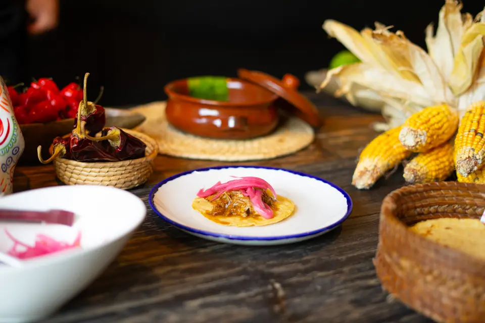 Small taco with pink pickled onion on a blue-rimmed plate, with corn cobs and dried chilies behind