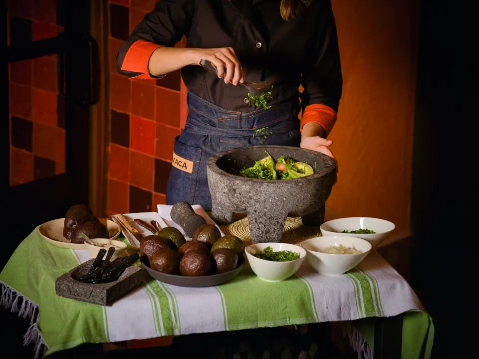 Person in a denim apron preparing guacamole in a stone mortar, with avocados and bowls in front