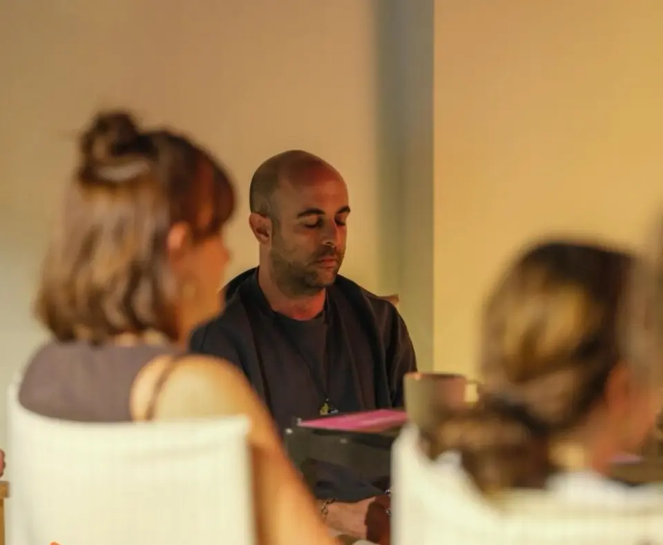 Person with closed eyes at a table, between two blurred people in the foreground, in warm light