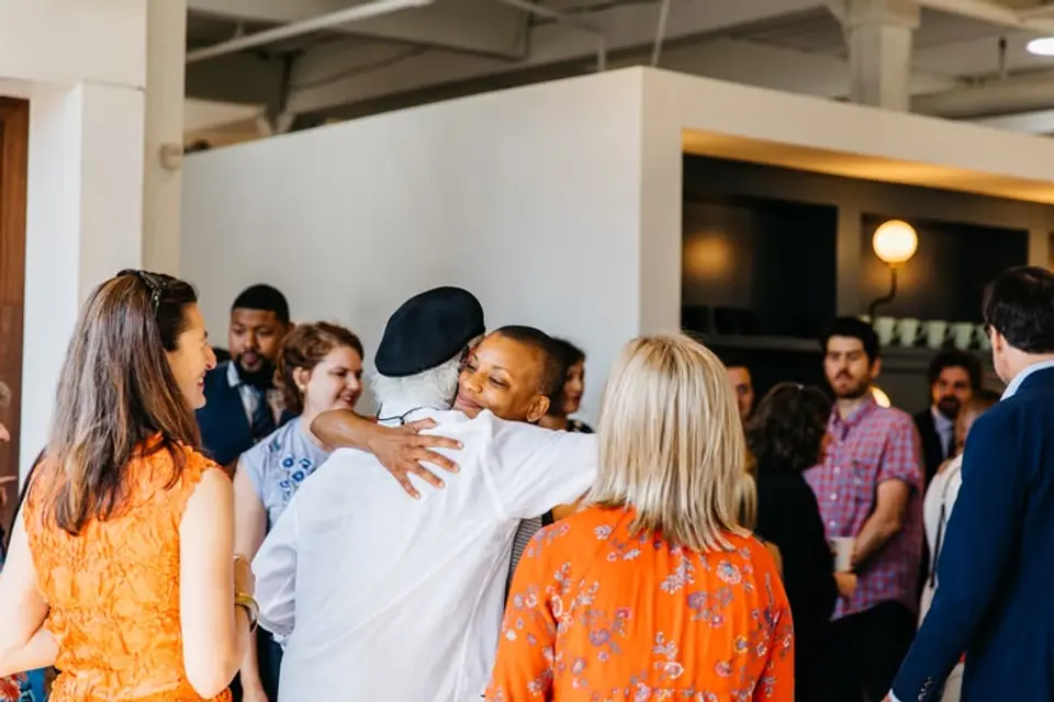 Two people hugging in a bright room, surrounded by standing guests in conversation