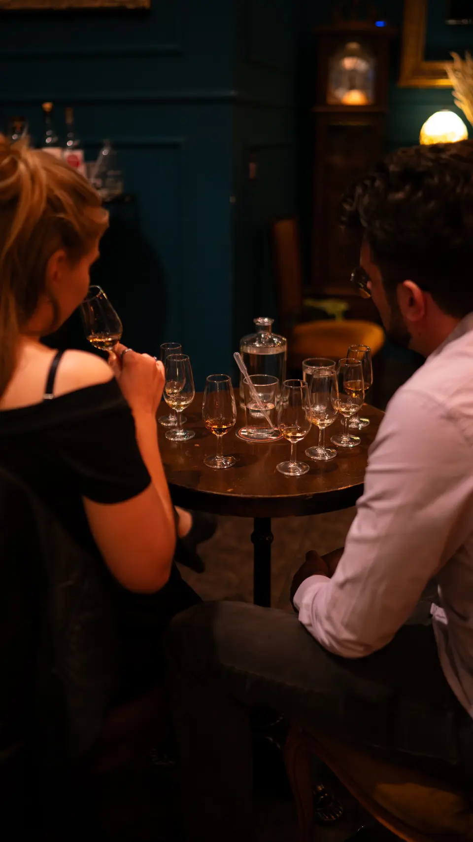 Two people seen from behind at a round table with tasting glasses, in a corner with dark blue walls