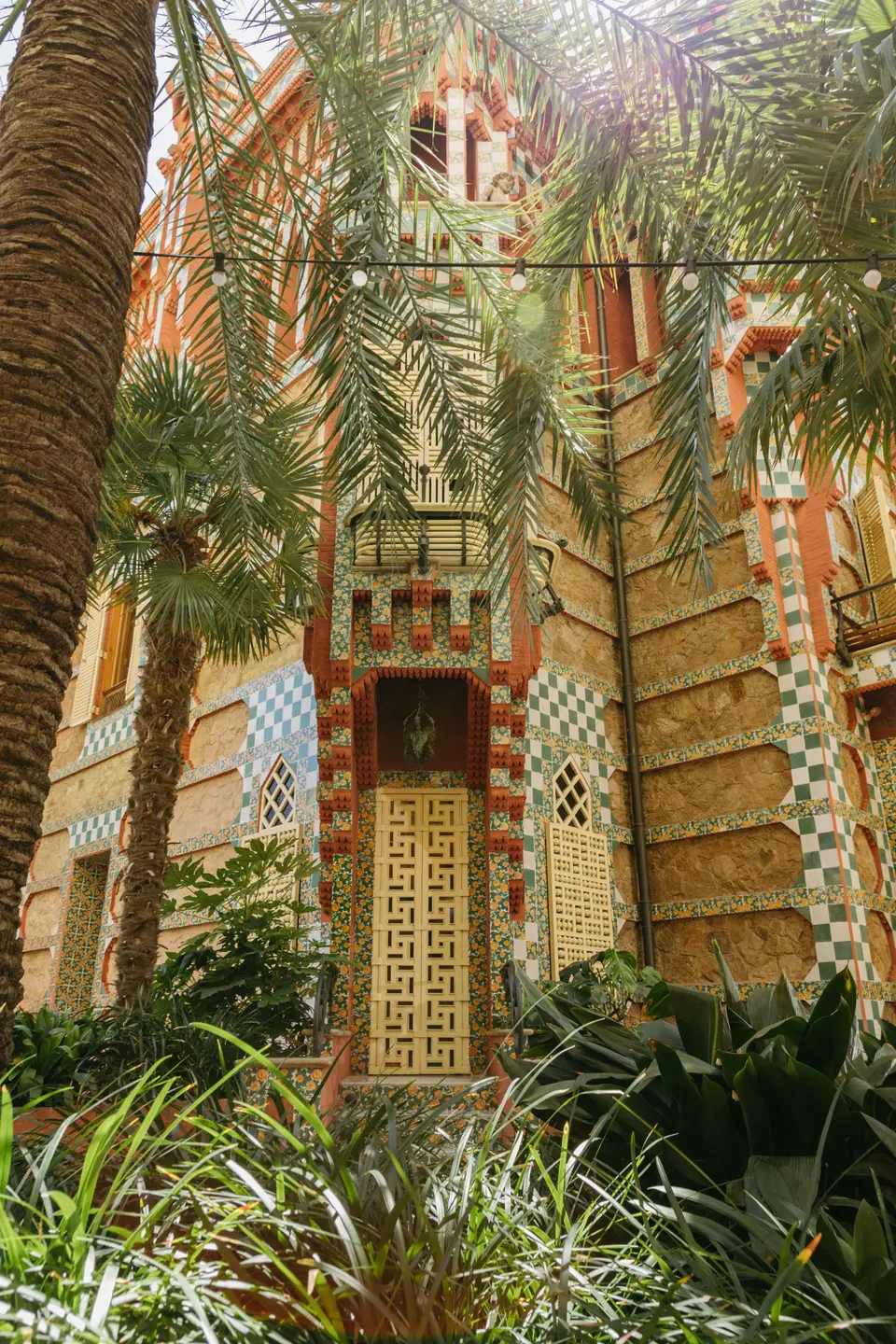 Façade with a golden lattice door seen between palms and plants, with sunlight coming through the leaves