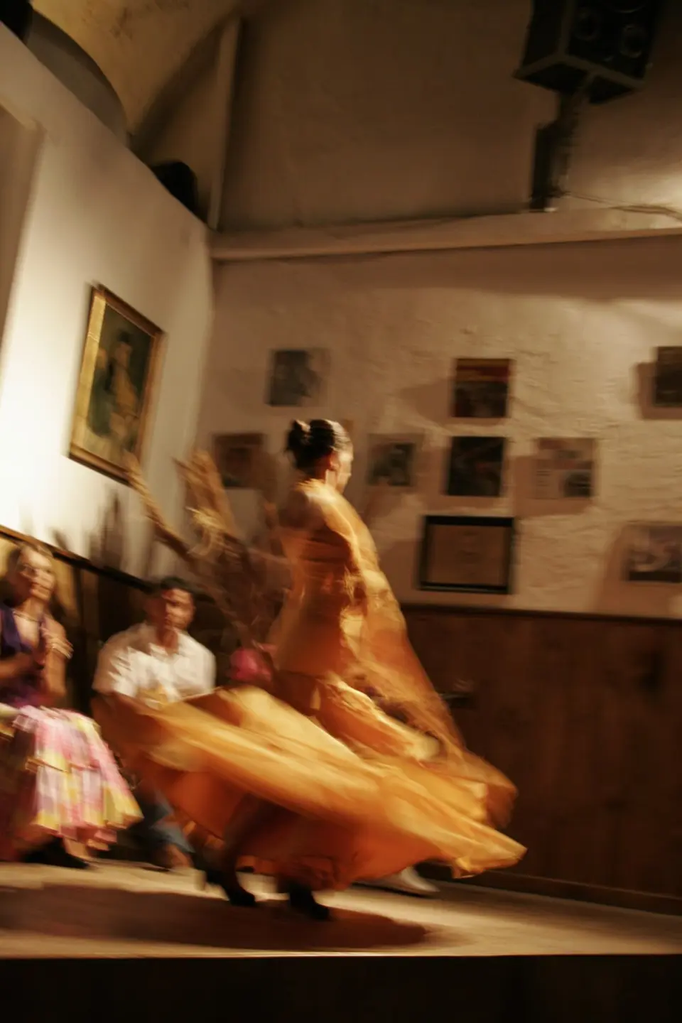 Dancer in an orange dress mid-spin on a small stage, with seated spectators in a white-walled room