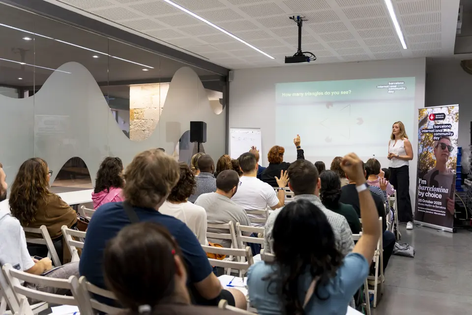 Attendees on folding chairs, some raising a hand, facing a projected screen in a modern room