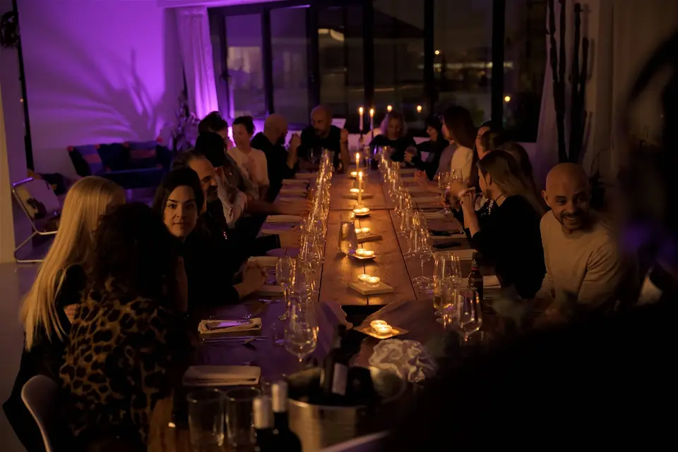 Long table with diners on both sides and lit candles, in a violet-lit room with large windows behind