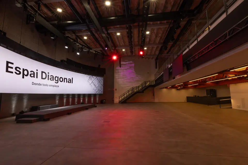 Empty main floor with a curved screen on the left, a low stage, a metal staircase at the back and spotlights overhead