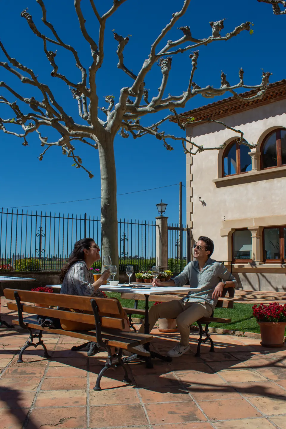 Two people seated at an outdoor table beside a stone building and a bare tree under a blue sky