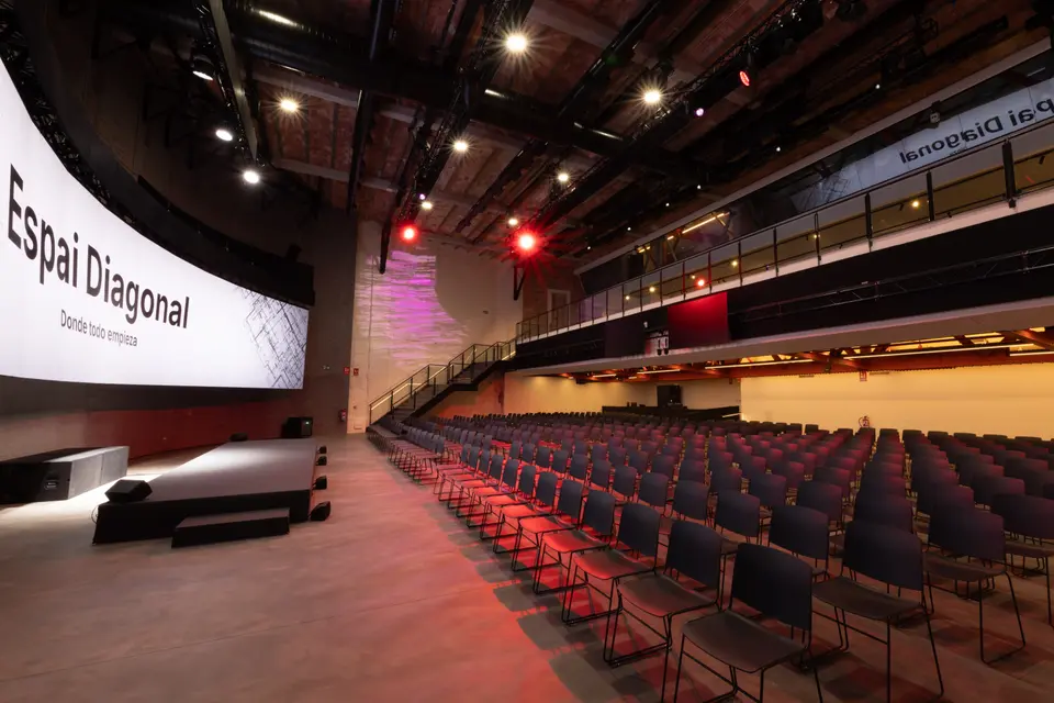 Theatre-style setup with hundreds of black chairs facing the stage and curved screen, with a raised gallery on the right