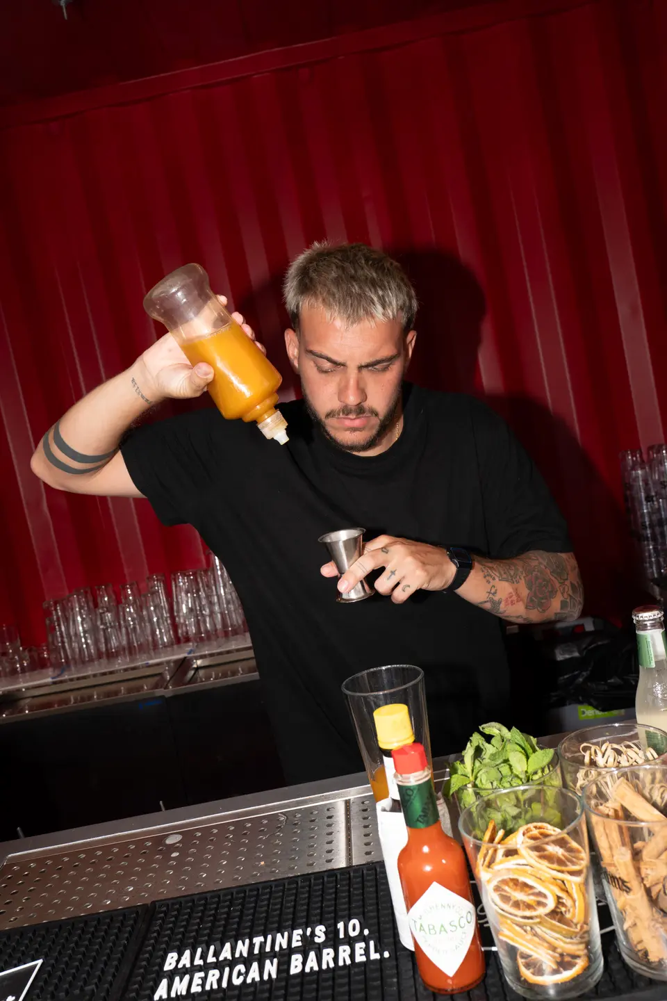 Bartender in a black T-shirt pouring orange syrup into a cocktail, with glasses and garnishes on a red container bar