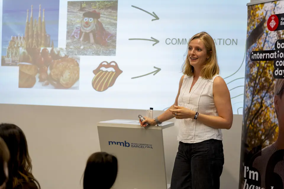 Speaker holding a clicker beside a lectern, with a projected presentation behind her and a roll-up banner