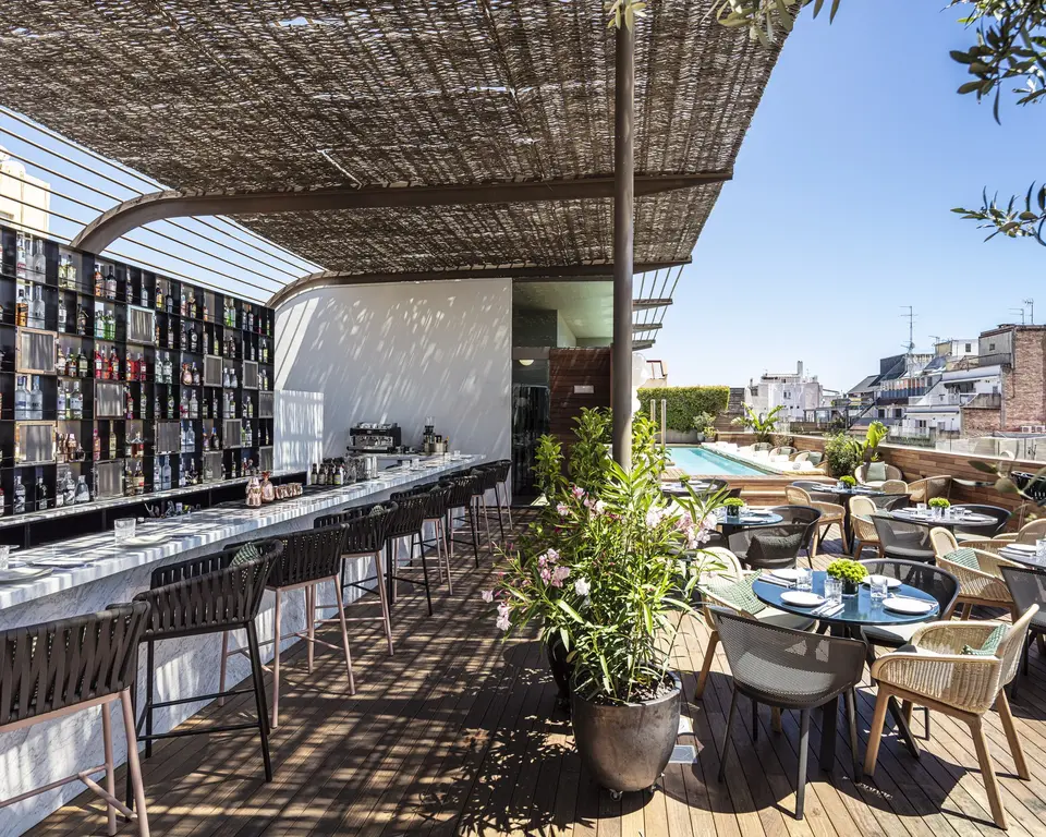 Wooden deck terrace with a marble bar and stools under a reed pergola, with the pool and dining tables beyond