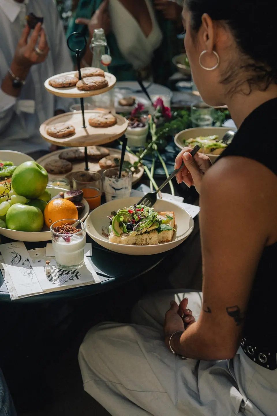 Brunch table with a tiered stand of cookies, fruit and plates of toast, and a person eating in the foreground