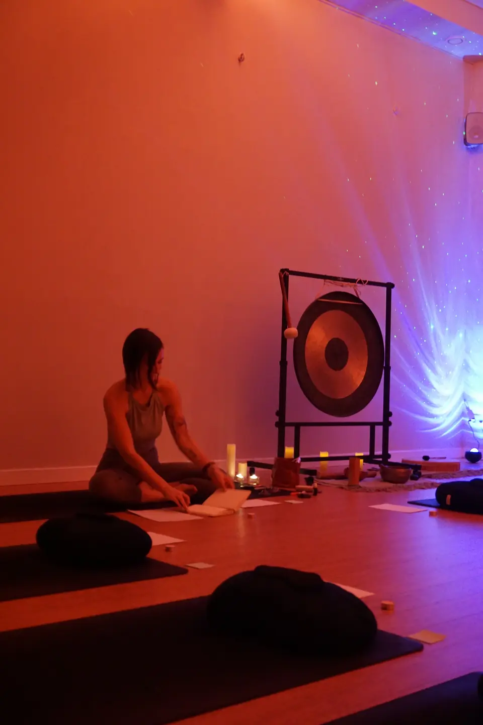 Person seated on the floor beside a gong and bowls, with mats and cushions in the foreground