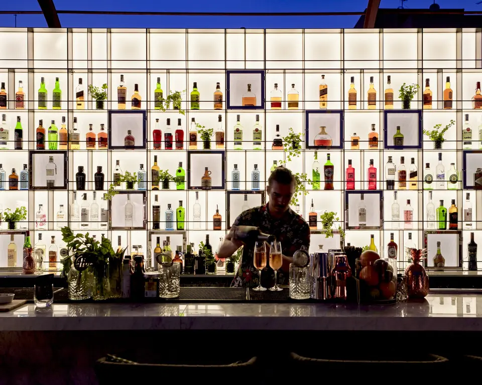 Bartender preparing cocktails at the bar, with bottles lit on a grid-pattern shelf wall at dusk