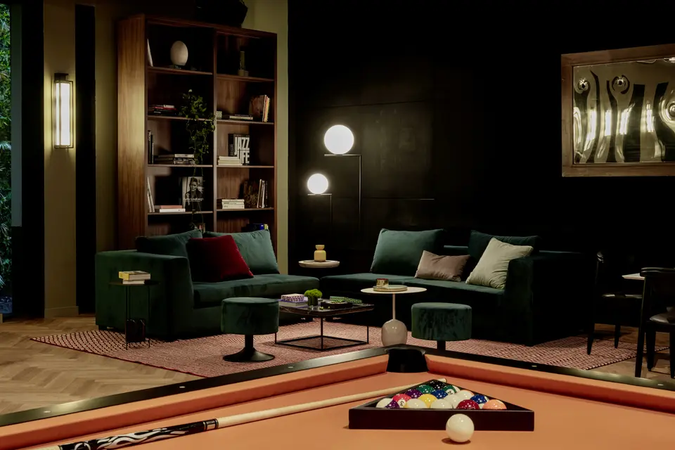 Lounge area with green sofas and a low table behind a pool table, with a wooden bookcase and dim lighting