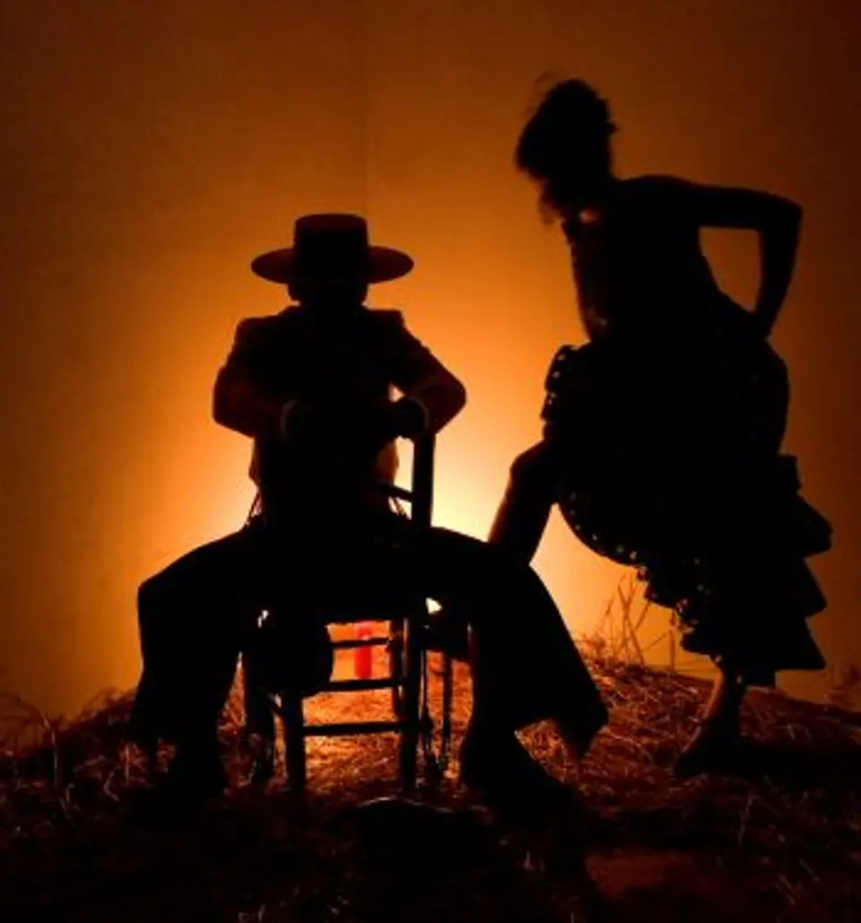Backlit silhouettes of a seated man in a hat and a dancer in a ruffled skirt against an orange glow