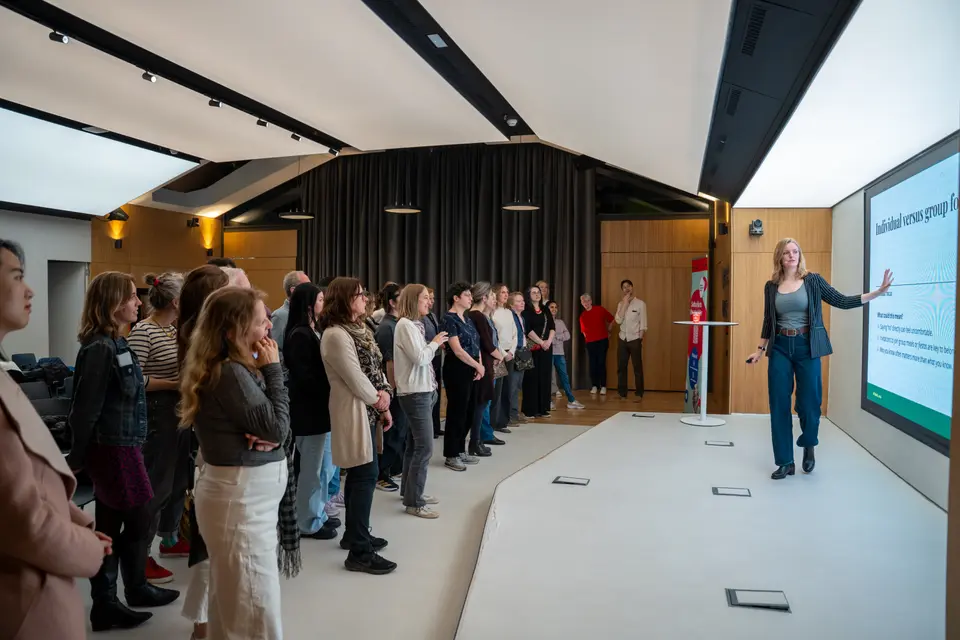 Attendees standing in an open hall watching a speaker point at a screen showing a presentation
