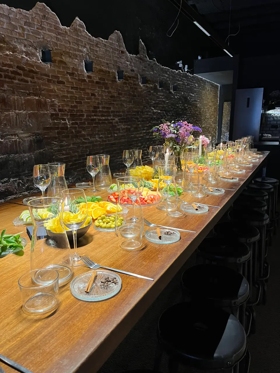 Long wooden table with cut fruit, glasses and jugs beside a brick wall, in a dim venue