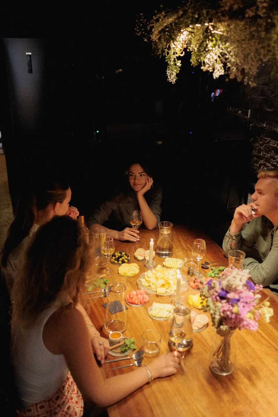 Several people seated at a wooden table with jugs, glasses and fruit platters by candlelight